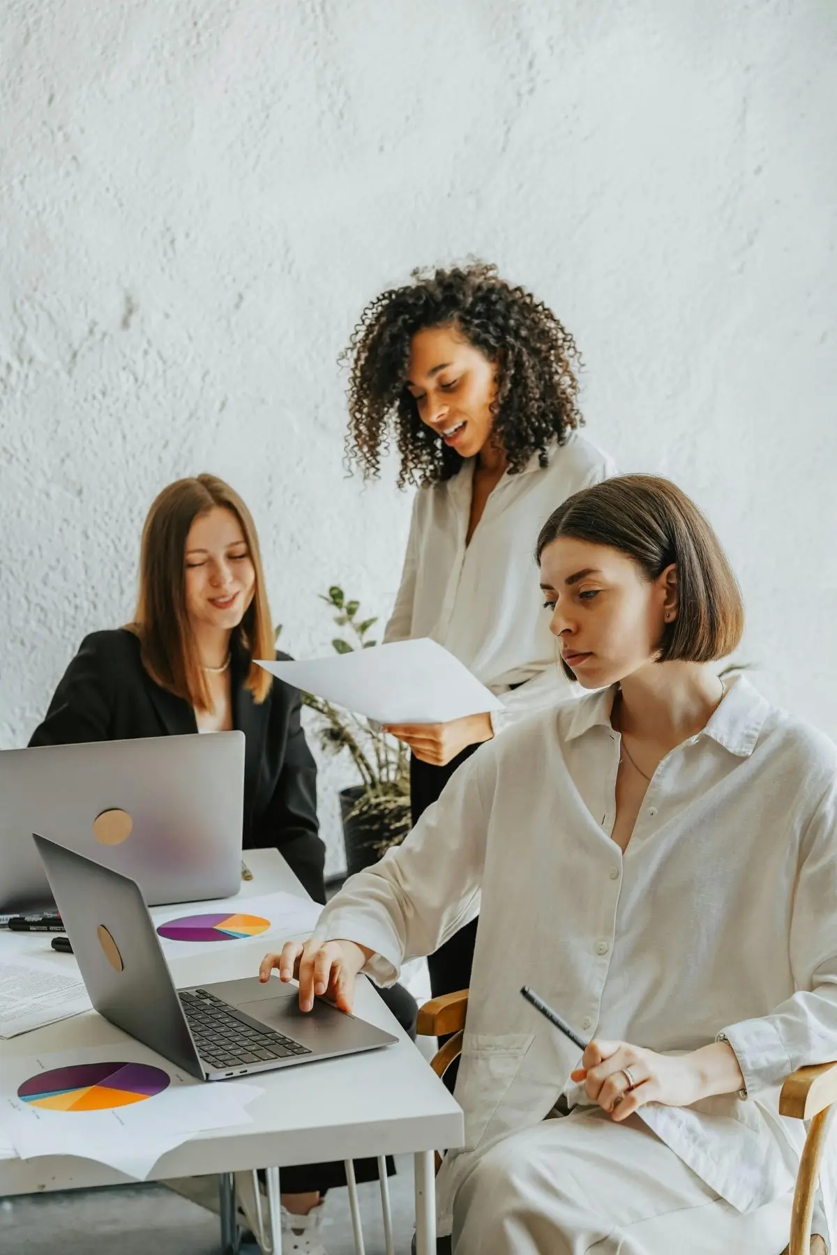 Three women working at a table with two laptops and papers, working on building their businesses